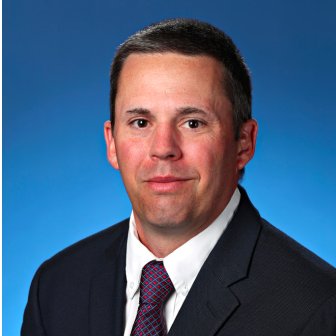 professional headshot of white male wearing a black suit jacket and red/blue tie against a blue background